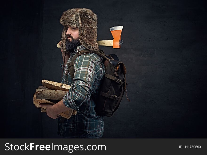 Brutal bearded middle age male dressed in a plaid shirt and a fur hat holds an axe and firewoods over dark grey background. Brutal bearded middle age male dressed in a plaid shirt and a fur hat holds an axe and firewoods over dark grey background.