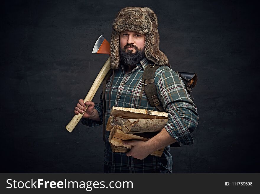 Brutal bearded middle age male dressed in a plaid shirt and a fur hat holds an axe and firewoods over dark grey background. Brutal bearded middle age male dressed in a plaid shirt and a fur hat holds an axe and firewoods over dark grey background.