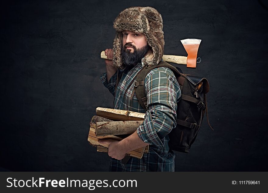 Brutal bearded middle age male dressed in a plaid shirt and a fur hat holds an axe and firewoods over dark grey background. Brutal bearded middle age male dressed in a plaid shirt and a fur hat holds an axe and firewoods over dark grey background.