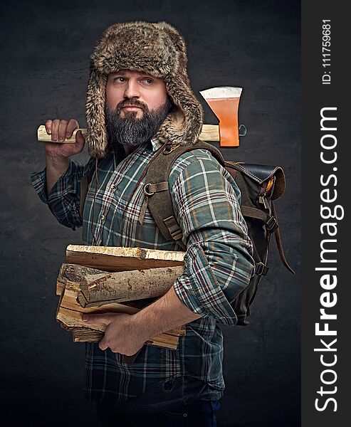 Brutal bearded middle age male dressed in a plaid shirt and a fur hat holds an axe and firewoods over dark grey background. Brutal bearded middle age male dressed in a plaid shirt and a fur hat holds an axe and firewoods over dark grey background.