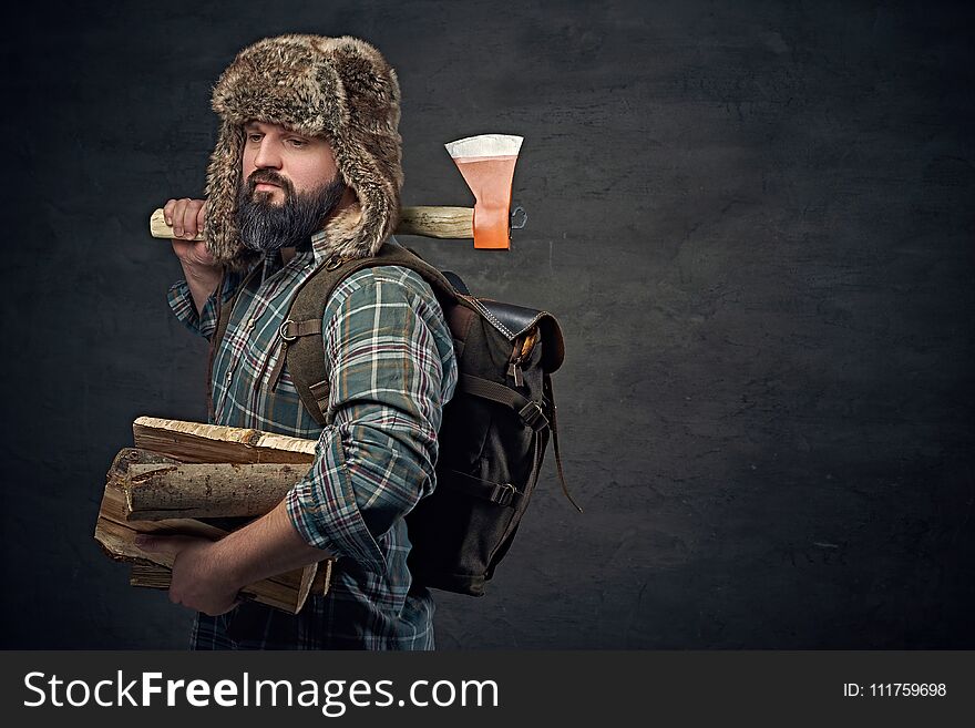 Brutal bearded middle age male dressed in a plaid shirt and a fur hat holds an axe and firewoods over dark grey background. Brutal bearded middle age male dressed in a plaid shirt and a fur hat holds an axe and firewoods over dark grey background.