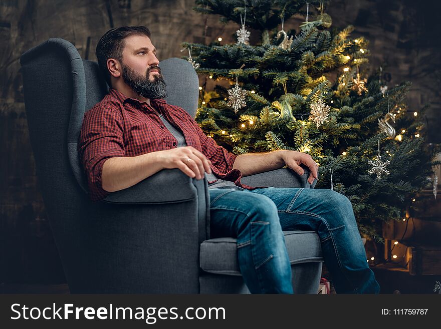 A bearded middle age male dressed in a plaid shirt and jeans sits on a chair over Christmas illumination and fir tree in background. A bearded middle age male dressed in a plaid shirt and jeans sits on a chair over Christmas illumination and fir tree in background.