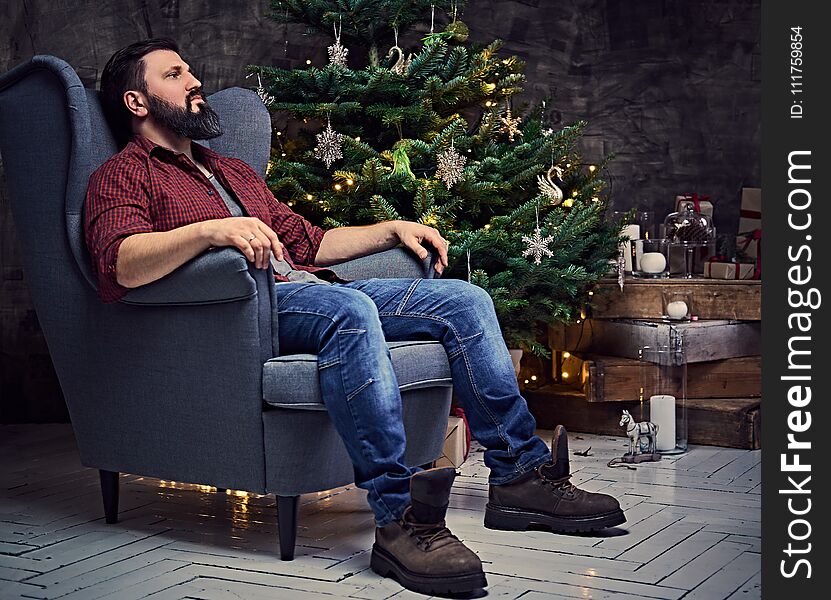 A bearded middle age male dressed in a plaid shirt and jeans sits on a chair over Christmas illumination and fir tree in background. A bearded middle age male dressed in a plaid shirt and jeans sits on a chair over Christmas illumination and fir tree in background.