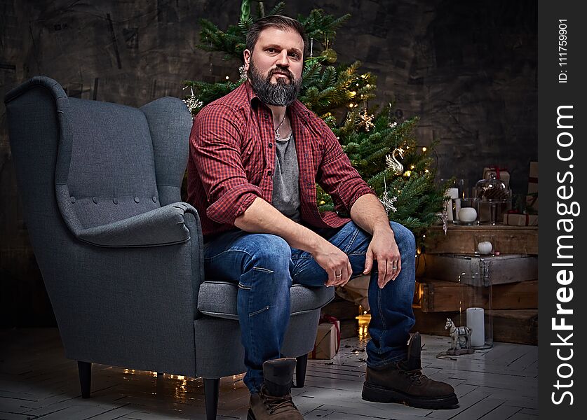 A bearded middle age male dressed in a plaid shirt and jeans sits on a chair over Christmas illumination and fir tree in background. A bearded middle age male dressed in a plaid shirt and jeans sits on a chair over Christmas illumination and fir tree in background.
