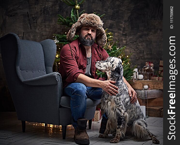 Bearded middle age male dressed in a plaid shirt and fur hat and Irish setter with fir tree and Christmas decoration on background. Bearded middle age male dressed in a plaid shirt and fur hat and Irish setter with fir tree and Christmas decoration on background.