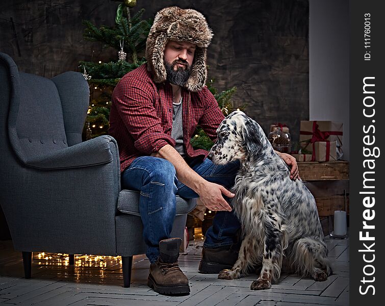 Bearded middle age male dressed in a plaid shirt and fur hat and two Irish setter dogs with fir tree and Christmas decoration on background. Bearded middle age male dressed in a plaid shirt and fur hat and two Irish setter dogs with fir tree and Christmas decoration on background.