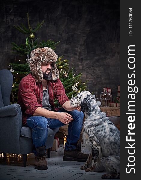 Bearded middle age male dressed in a plaid shirt and fur hat and two Irish setter dogs with fir tree and Christmas decoration on background. Bearded middle age male dressed in a plaid shirt and fur hat and two Irish setter dogs with fir tree and Christmas decoration on background.