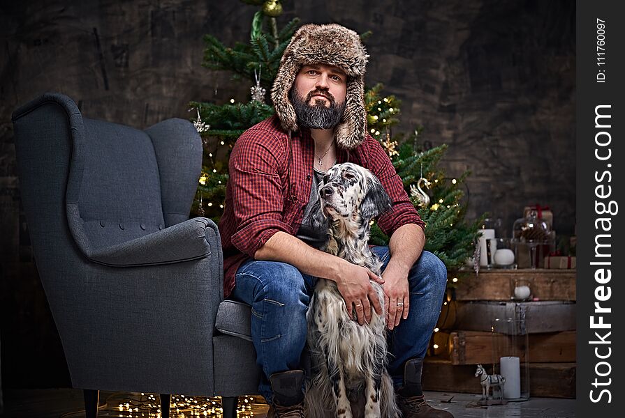 Bearded middle age male dressed in a plaid shirt and fur hat and two Irish setter dogs with fir tree and Christmas decoration on background. Bearded middle age male dressed in a plaid shirt and fur hat and two Irish setter dogs with fir tree and Christmas decoration on background.