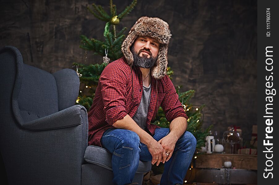 Bearded middle age male dressed in plaid shirt, jeans and fur hat sits on a chair over Christmas illumination and fir tree on background. Bearded middle age male dressed in plaid shirt, jeans and fur hat sits on a chair over Christmas illumination and fir tree on background.