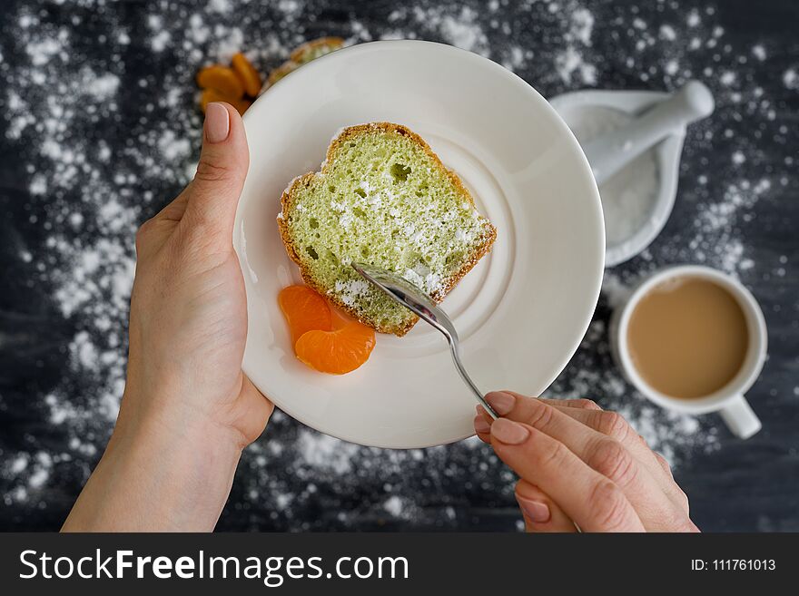 Mint cake on a plate with tangerines in hands. The background is a dark surface sprinkled with powdered sugar, a cup of coffee.