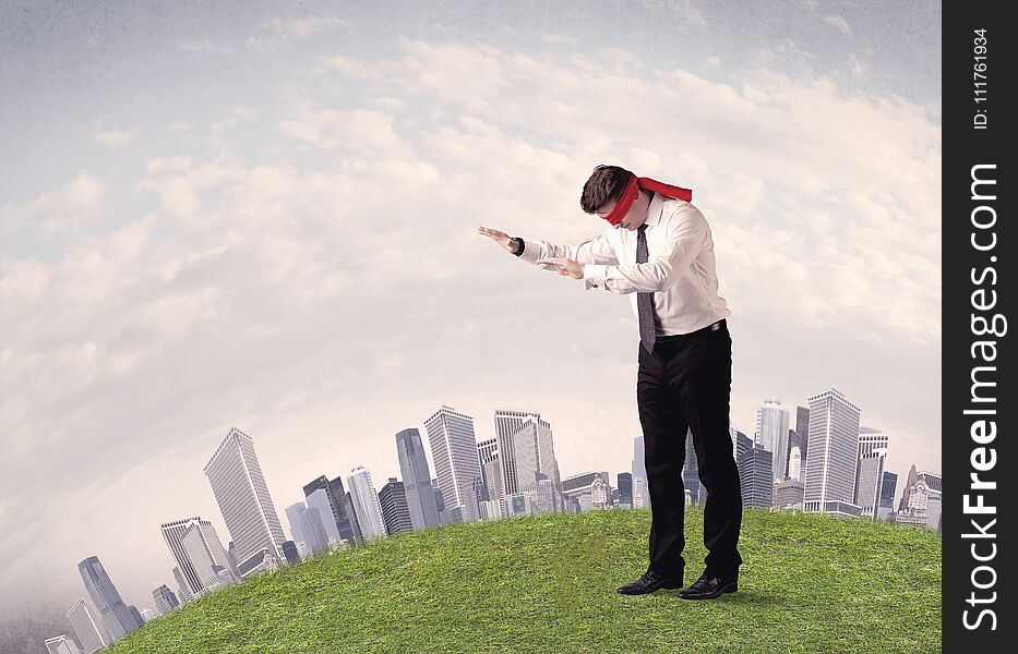 Young blindfolded businessman steps on a a patch of grass with a grey buildings in the background. Young blindfolded businessman steps on a a patch of grass with a grey buildings in the background