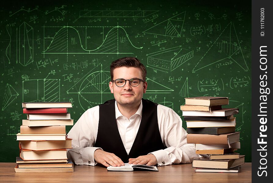 A young passionate male teacher sitting at school desk, reading a book, with area algorythm calculations and numbers on the blackboard concept. A young passionate male teacher sitting at school desk, reading a book, with area algorythm calculations and numbers on the blackboard concept.