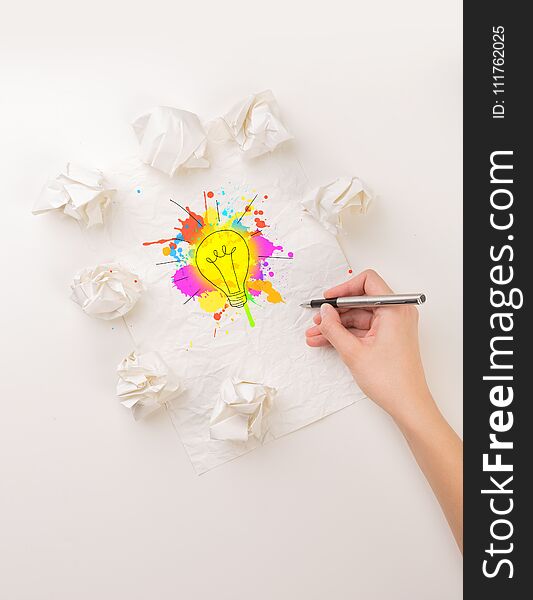 Female hand next to a few crumpled paper balls drawing a colorful lightbulb. Female hand next to a few crumpled paper balls drawing a colorful lightbulb