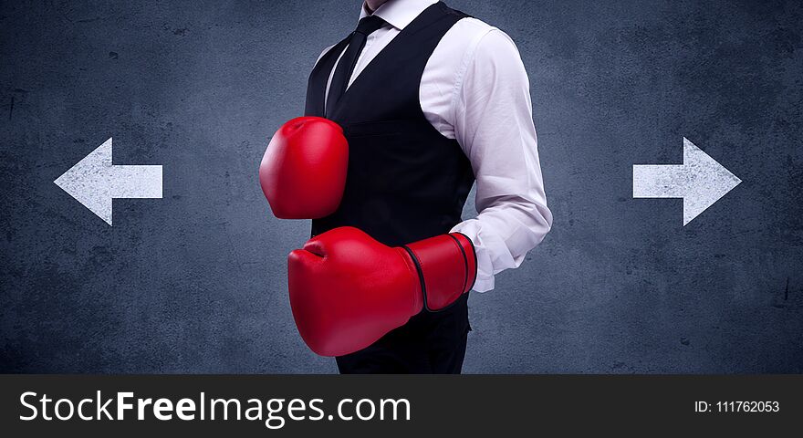 A confident businessman standing with red boxing gloves on his hand in front of arrows pointing in different directions on urban wall background concept. A confident businessman standing with red boxing gloves on his hand in front of arrows pointing in different directions on urban wall background concept.