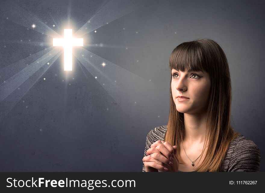 Young woman praying on a grey background with a shiny cross above her. Young woman praying on a grey background with a shiny cross above her