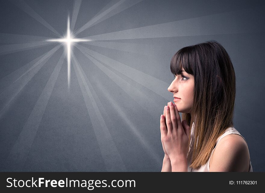 Young woman praying on a grey background with a shiny cross silhouette above her. Young woman praying on a grey background with a shiny cross silhouette above her