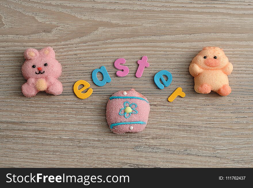 A pink bunny shape marshmallow, a chicken shape marshmallow and an egg with the word easter on a wooden background