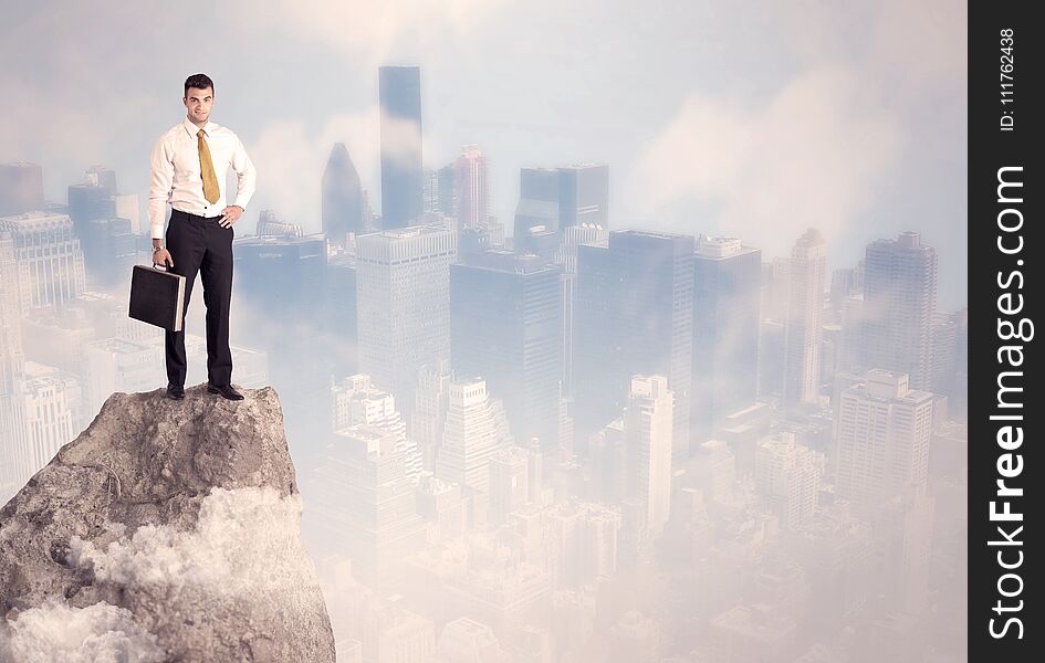 A successful good looking businessman standing on top of a high cliff above the city scape with clouds concept. A successful good looking businessman standing on top of a high cliff above the city scape with clouds concept