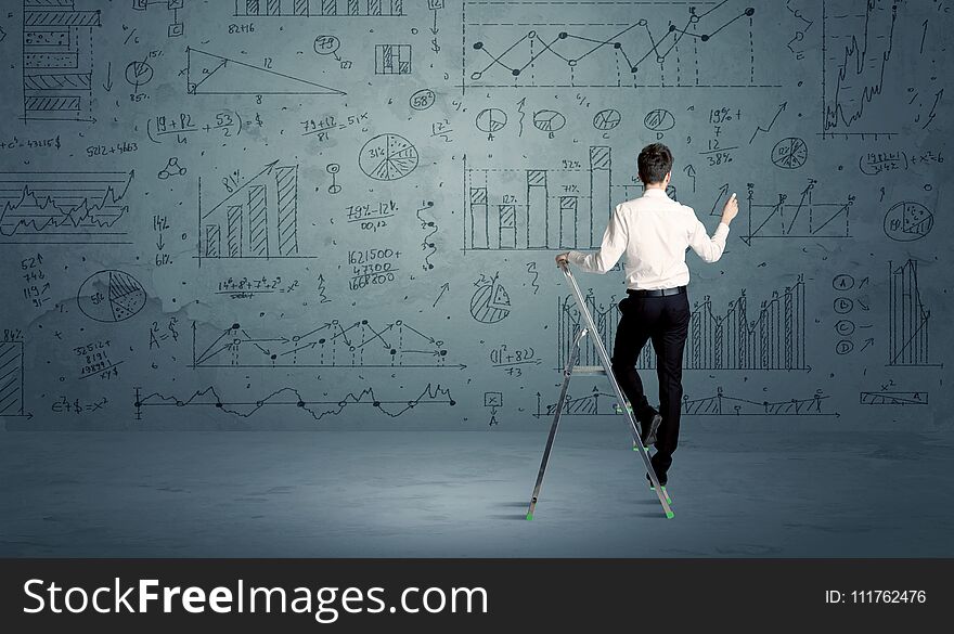 A businessman in modern stylish elegant suit standing on a small ladder and drawing pie and block charts on grey wall background with exponential progressing curves, lines, circles, angles, blocks, numbers. A businessman in modern stylish elegant suit standing on a small ladder and drawing pie and block charts on grey wall background with exponential progressing curves, lines, circles, angles, blocks, numbers