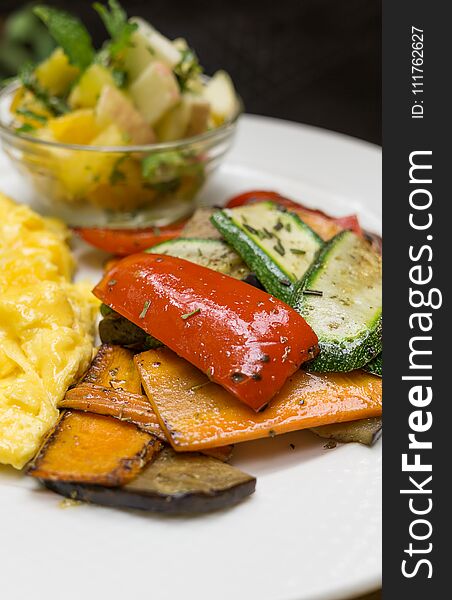Baked vegetables on white plate: sliced carrots, bell pepper, zucchini with scrambled eggs and blurred mango apple salad in backgr