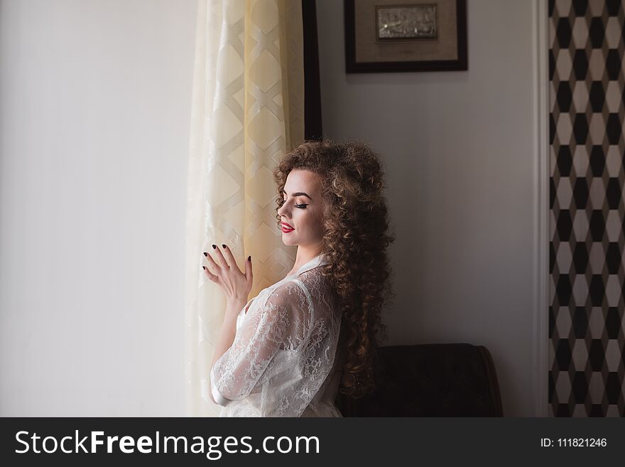 A happy girl stands by the window with her curly hair and is dressed in a white robe. A happy girl stands by the window with her curly hair and is dressed in a white robe