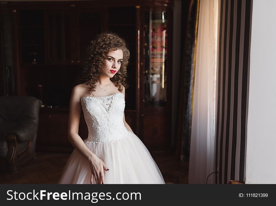 An elegant girl standing by the window dressed in a wedding dress she has curly hair