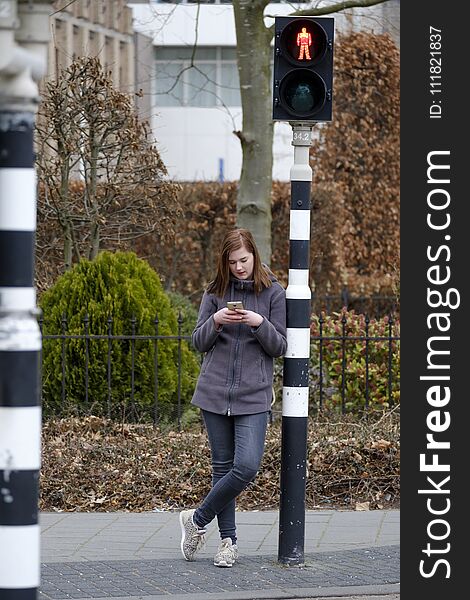 Young woman waits at a traffic light that is on red but does not pay attention to the traffic because she looks at her mobile phone. Young woman waits at a traffic light that is on red but does not pay attention to the traffic because she looks at her mobile phone.