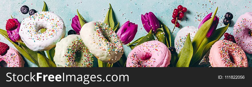 Sweet and colourful doughnuts with sprinkles, purple tulips and berries falling or flying in motion against blue pastel background. Border with space for text
