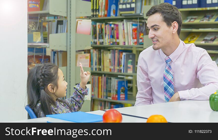 Smiley caucasian teacher and grouping of asian kids student learning and talking at white table and color book with bookshelf background