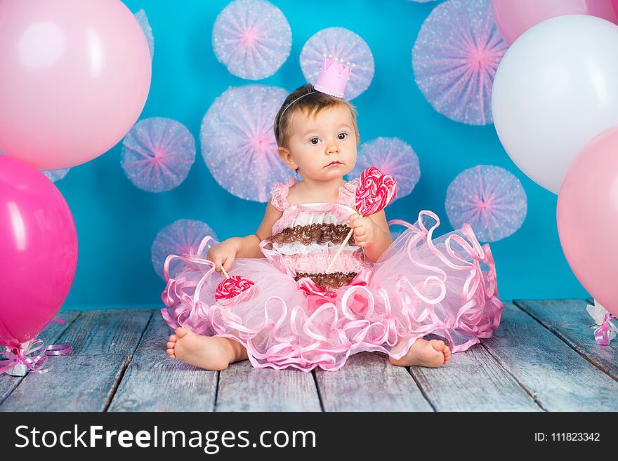 Funny child with a lollipop in the shape of a heart, happy little girl with a crown on her head eating a large sugar lollipop, isolated on a bright blue background with pink balls, studio. Funny child with a lollipop in the shape of a heart, happy little girl with a crown on her head eating a large sugar lollipop, isolated on a bright blue background with pink balls, studio