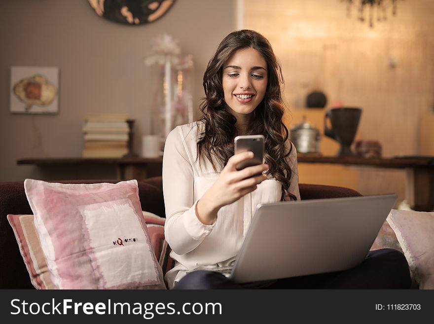 Woman Sitting On Sofa While Looking At Phone With Laptop On Lap