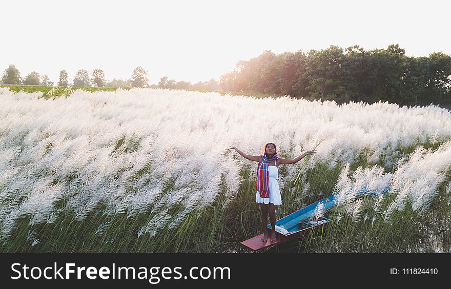 Portrait of beautiful Asian woman standing on fishing boat, enjoy natural outdoor at lake with grass flower at sunset
