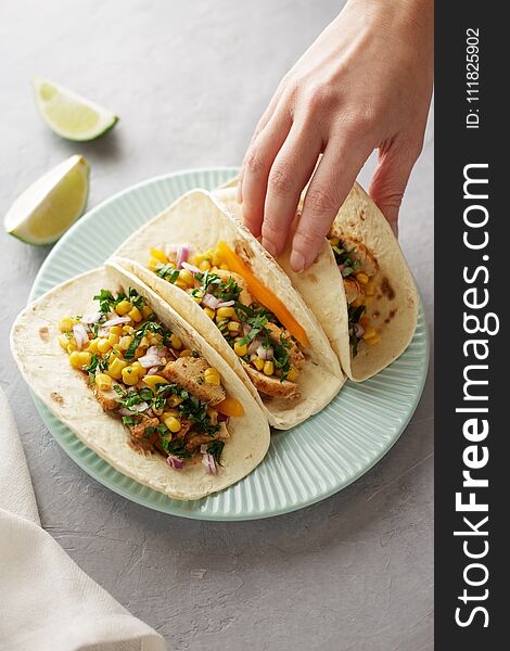 Hand takes taco from the blue plate on the dinner table. Mexican snacks with flatbread, corn, chicken meat and parsley.