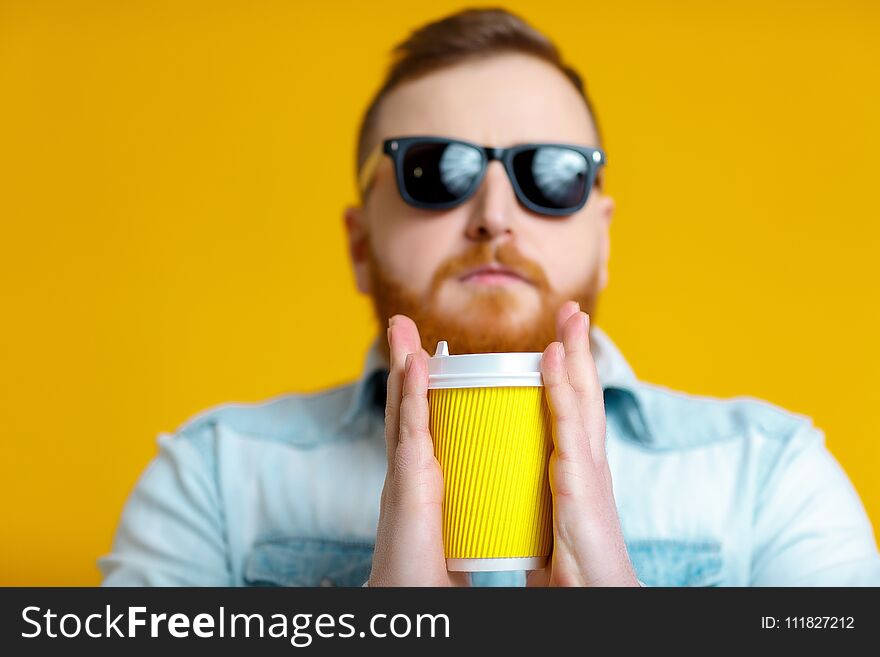 Red beard man in sunglasses and denim shirt holding paper cup with coffee on yellow background. Red beard man in sunglasses and denim shirt holding paper cup with coffee on yellow background.