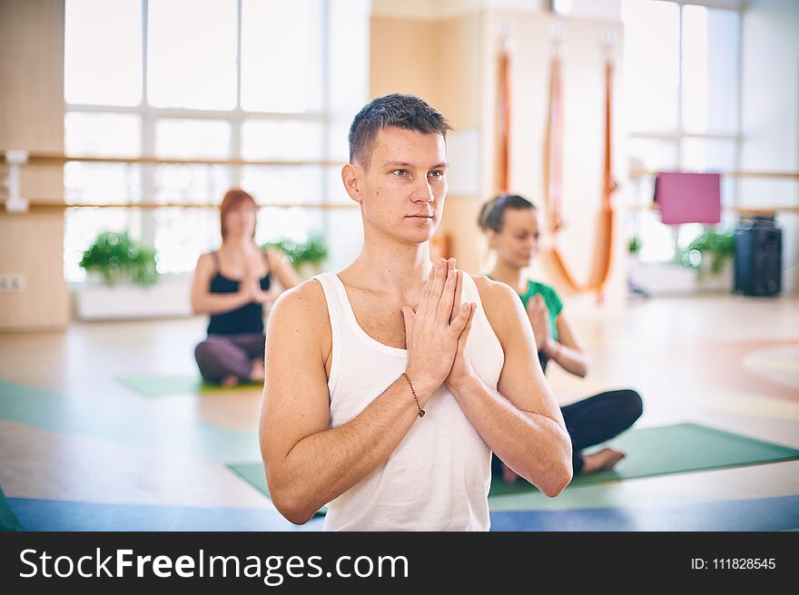 Group of young sporty people practicing yoga lesson with instructor, sitting in Padmasana exercise, Lotus pose with folded hands in namaste in yoga studio