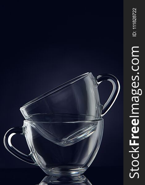 Conceptual composition of two glass teacups over dark grey background, close-up, vertical.