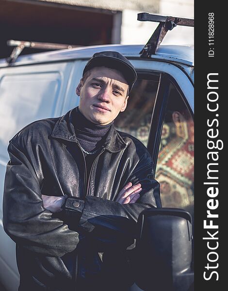 A young guy of criminal appearance in a black leather jacket stands near an old white van.