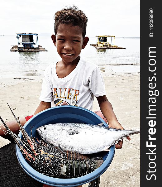 Boy Wearing White Crew-neck T-shirt Holding Blue Plastic Basin Full Of Lobster And Fish