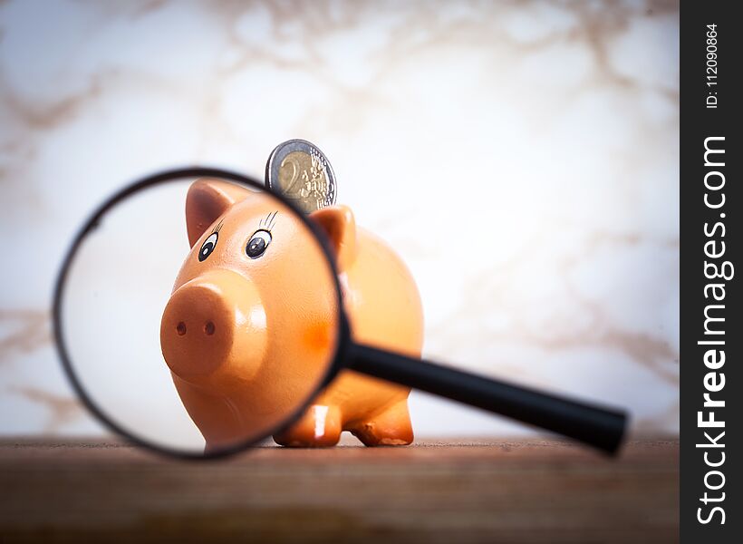 Piggy bank holding two euro coin and looking into magnifying glass. Piggy bank holding two euro coin and looking into magnifying glass