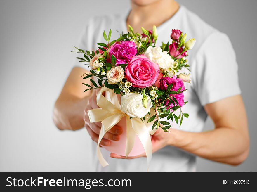 Man in white t shirt holding in hand rich gift bouquet. Composition of flowers in a pink hatbox. Tied with wide white ribbon and bow.