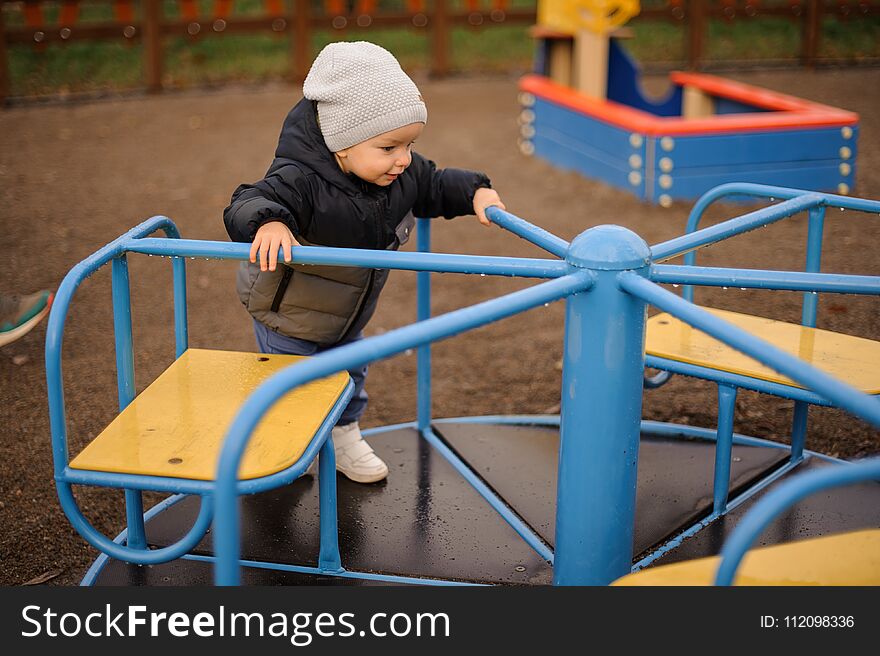 Cute little boy dressed in a warm hat and jacket riding on the carousel on the playground