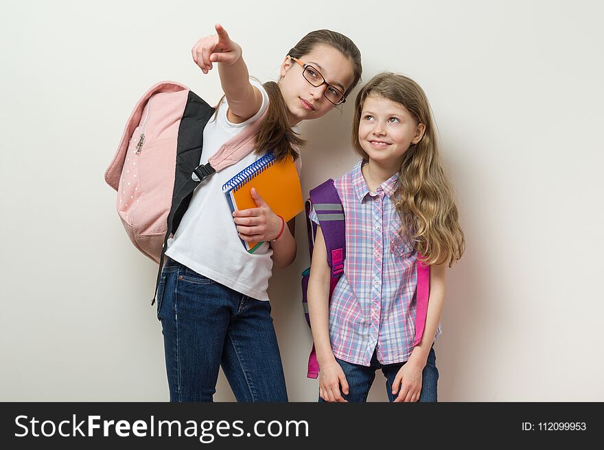 Two schoolchildren stand with backpacks background bright wall in the school, look and show to the side