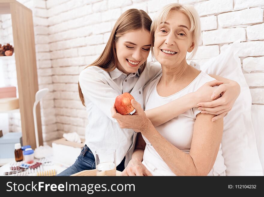 Girl is nursing elderly women in bed at home. They are embracing. Woman is holding apple.