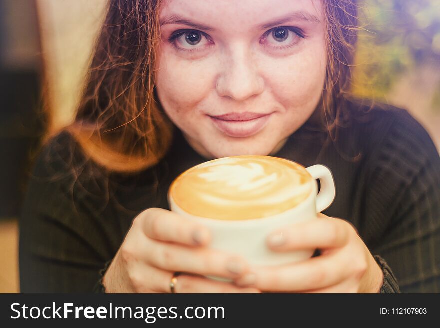 Beautiful girl with red hair and freckles smiles and drinks his coffee, cappuccino from the coffee Cup, portrait. Beautiful girl with red hair and freckles smiles and drinks his coffee, cappuccino from the coffee Cup, portrait