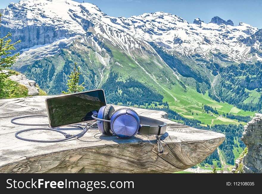 Cell phone with headphones on a wooden table on the background of the Swiss Alps and mountain scenery.