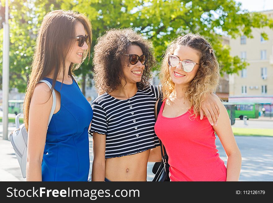 Outdoors portrait of three happy female multiethnic friends in sunglasses. Girls in colourful clothes having a walk in city in summer and having fun, copy space. Urban lifestyle, friendship concept. Outdoors portrait of three happy female multiethnic friends in sunglasses. Girls in colourful clothes having a walk in city in summer and having fun, copy space. Urban lifestyle, friendship concept