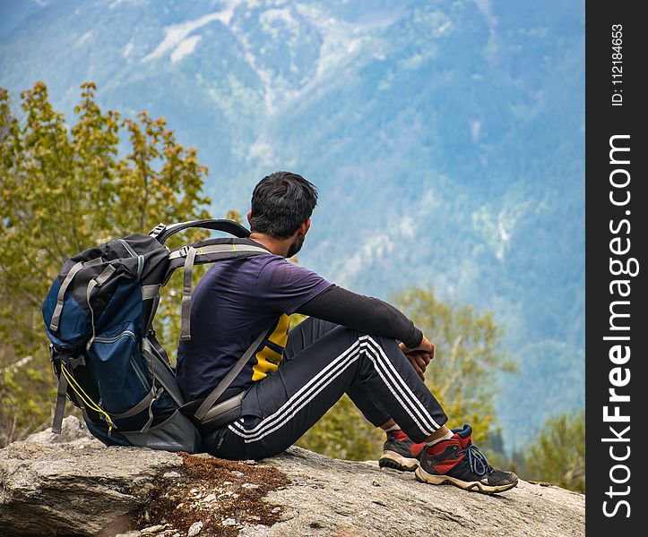 Man in Purple T-shirt With Blue Backpack Sitting on Gray Boulder