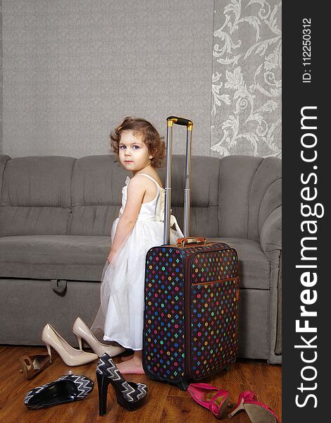 The little girl in a white dress with a suitcase beside her and the adult women`s shoes. The little girl in a white dress with a suitcase beside her and the adult women`s shoes.