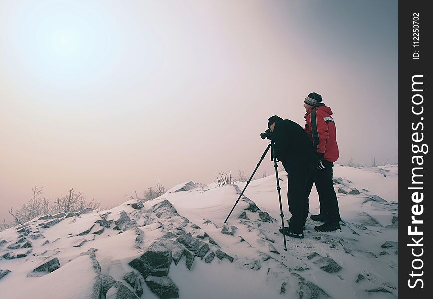 Hiker and photo enthusiast stay on snowy peak at tripod. Men on cliff speaking and thinking. Dreamy winter landscape with misty sunrise