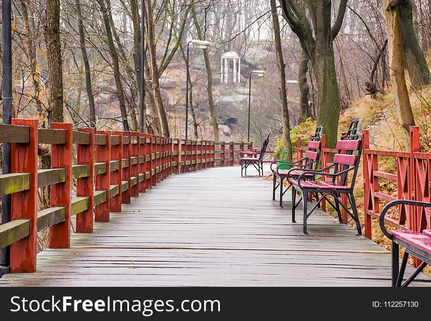 Wooden path - hinged bridge with wet boards, wrought iron benches and lanterns in the park on a hillside overlooking the white rotunda and granite stones in the background. Autumn rainy wet landscape